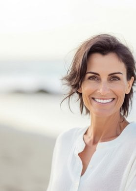 Een lachende vrouw met kort, donkerbruin haar, gekleed in een witte blouse, poseert op een zonnig strand met de zee op de achtergrond.