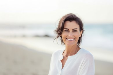 Een lachende vrouw met kort, donkerbruin haar, gekleed in een witte blouse, poseert op een zonnig strand met de zee op de achtergrond.